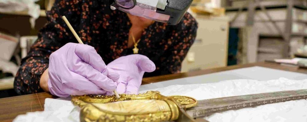 Restauratorin untersucht ein vergoldetes Museumsobjekt mit Lupe und Handschuhen
